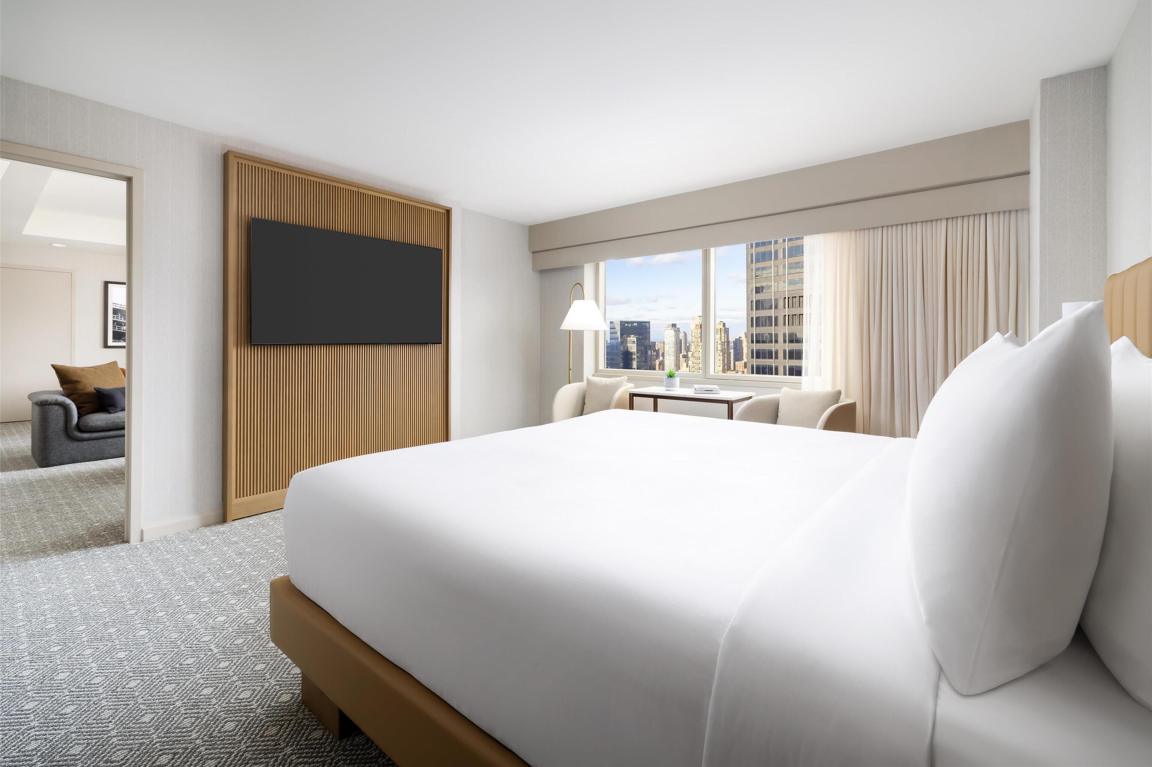 Habitación de hotel con una cama grande, un escritorio, una silla, un televisor montado en la pared y vistas de la ciudad a través del ventanal.