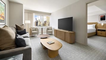 Quarto de hotel moderno com sofá, mesa redonda pequena, televisão de tela plana, cama e vista para o horizonte urbano.