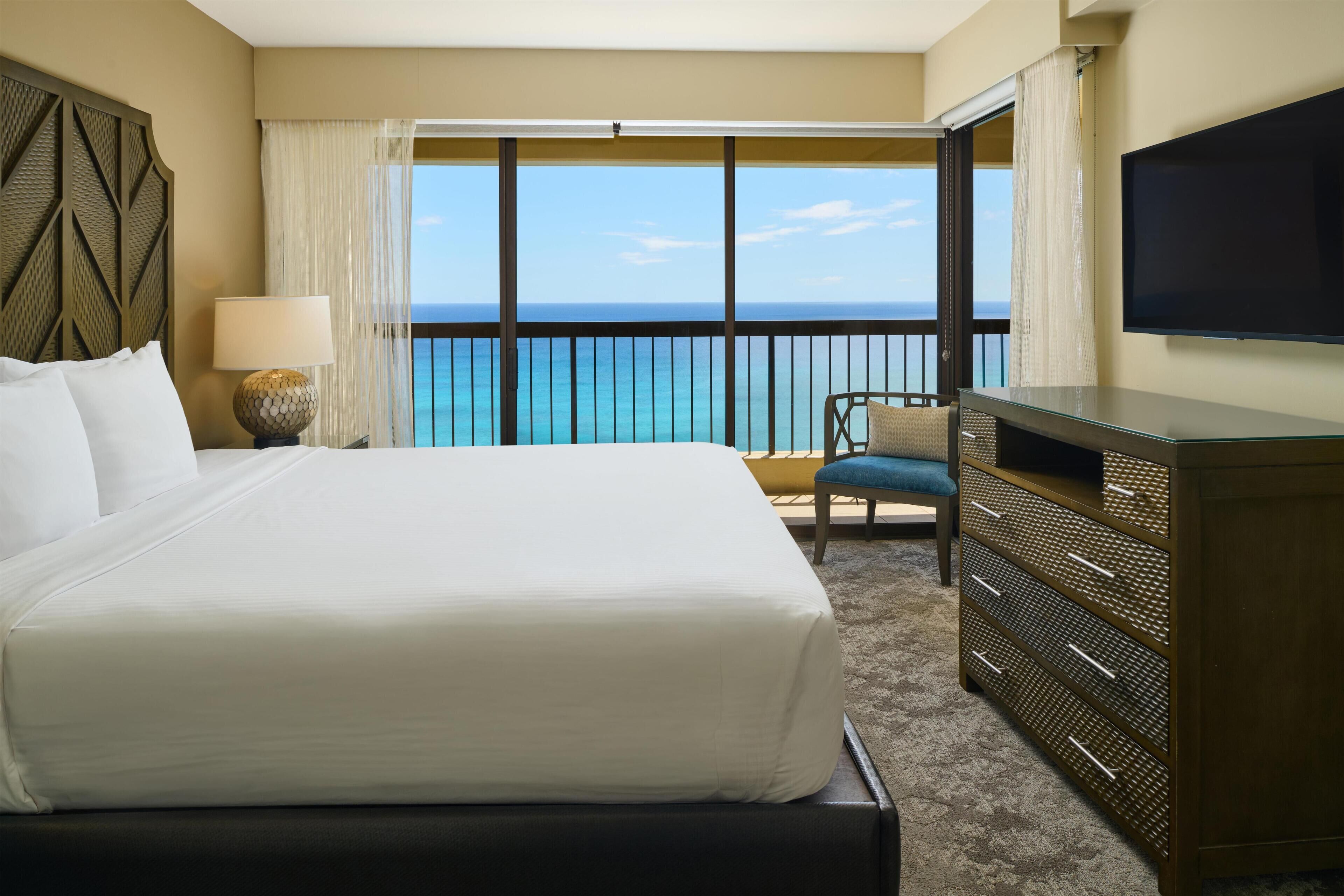 A hotel room with a large bed, a television, a chair, and a view of the ocean.