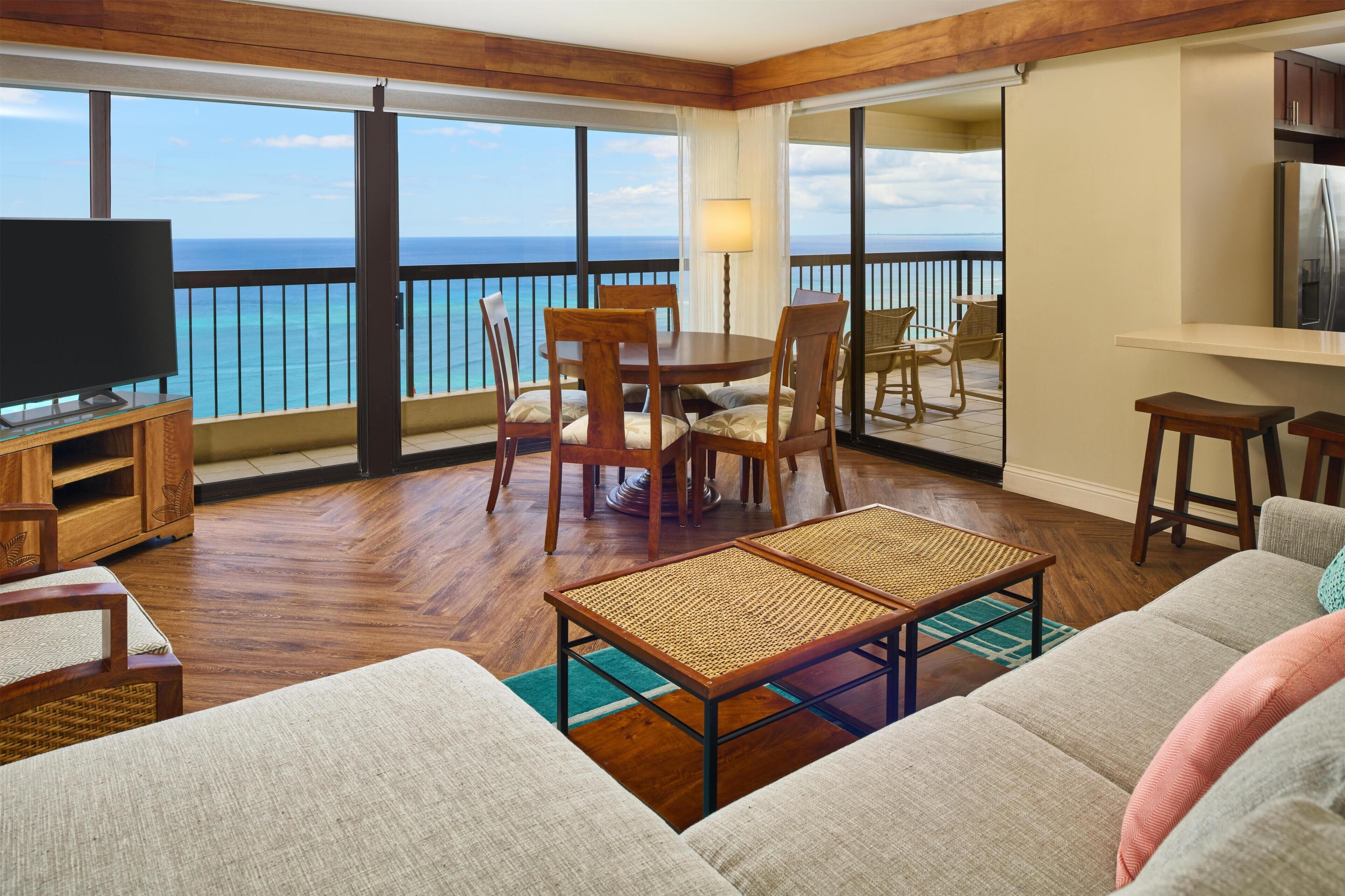 A spacious living area with a large window overlooking the ocean, furnished with a sofa, chairs, a coffee table, and a television.