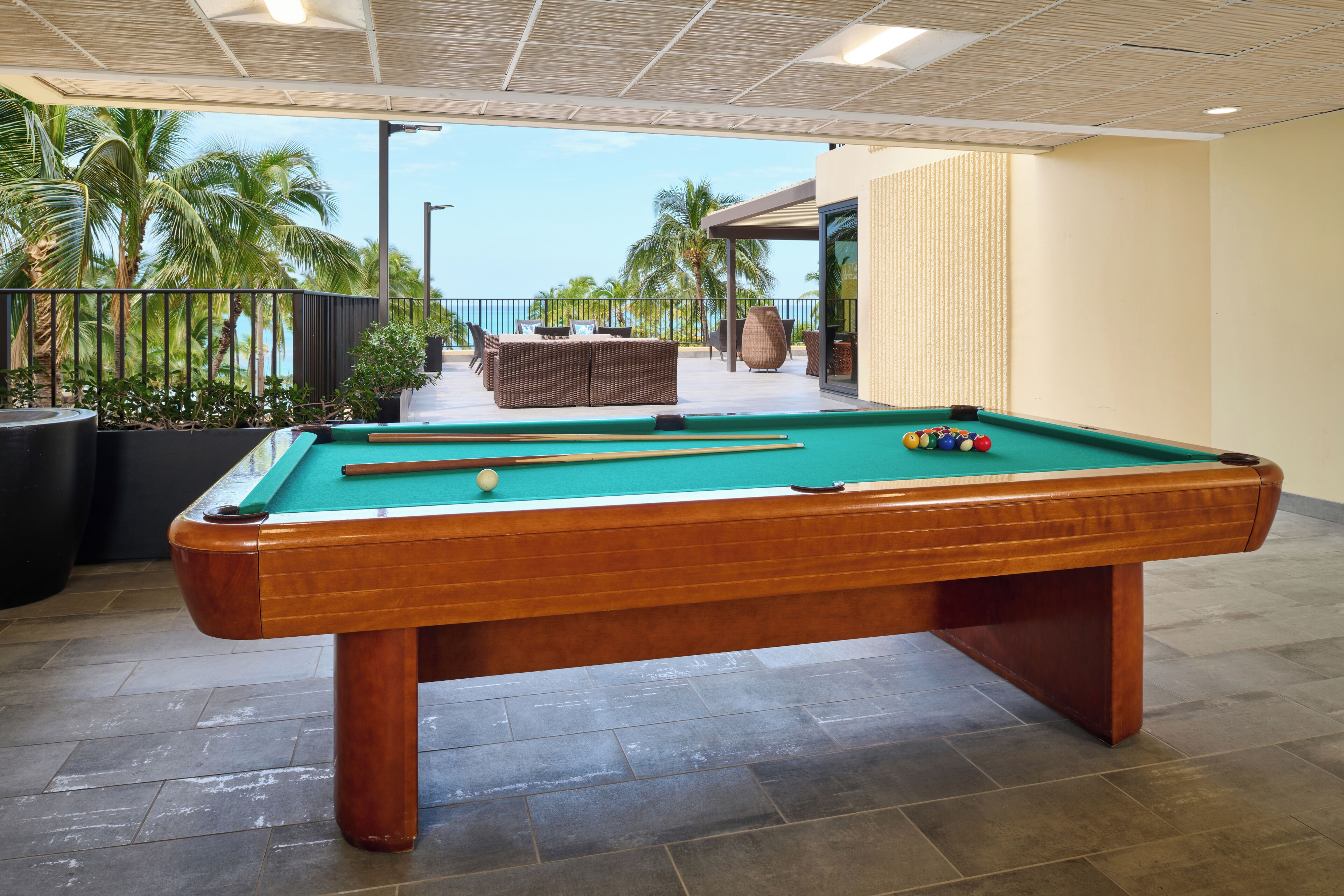A pool table with a green felt surface, surrounded by a tiled floor, and a view of palm trees and a building in the background.