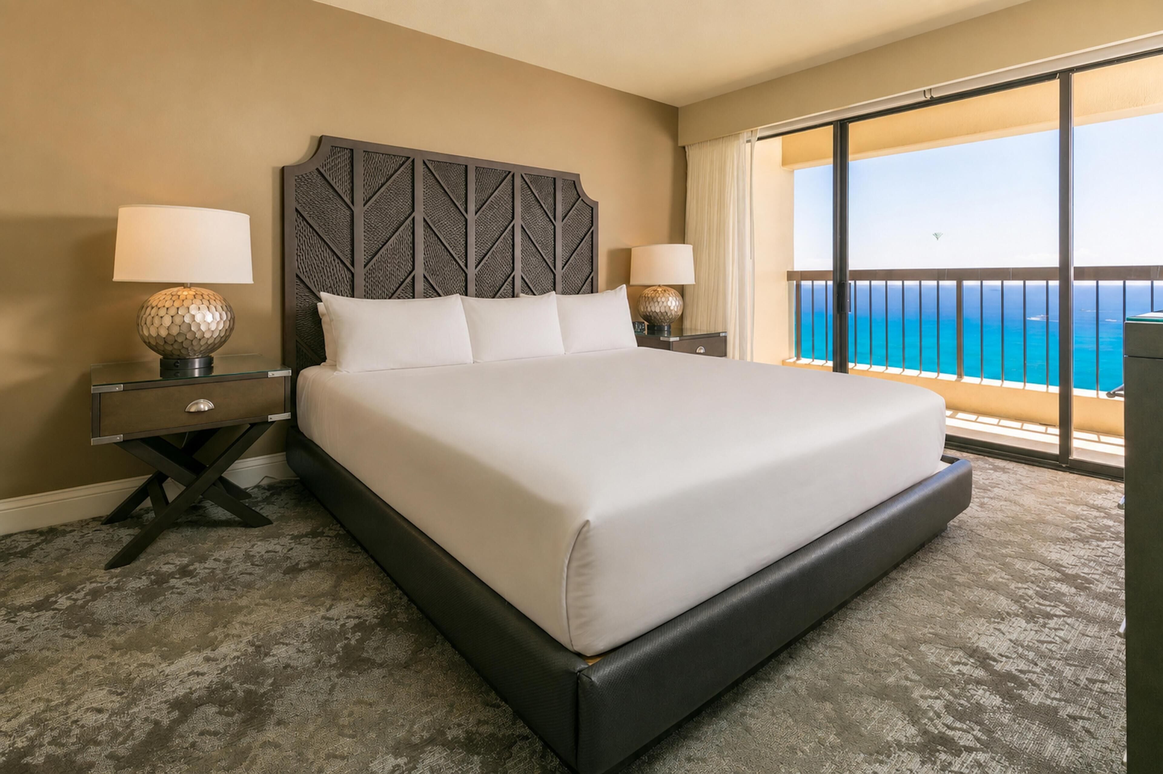 A hotel room with a large bed, two bedside tables with lamps, a view of the ocean through a balcony.
