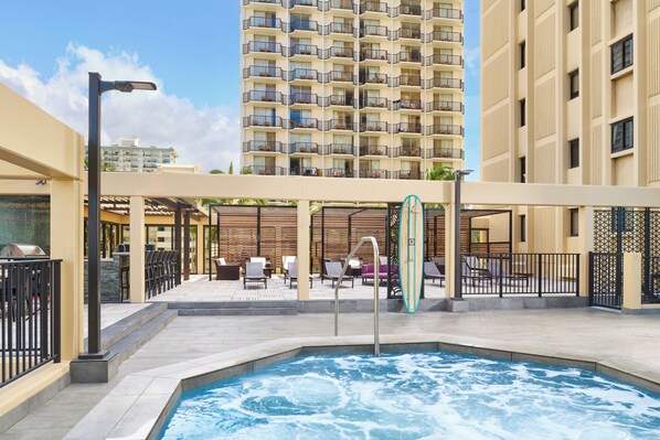 A rooftop pool area with lounge chairs and a surfboard, surrounded by high-rise buildings. - Aston Waikiki Beach Tower (Honolulu)