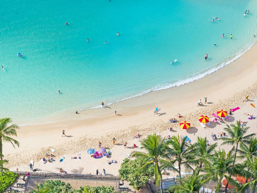 Una playa con agua turquesa cristalina, palmeras y gente tomando el sol y nadando.
