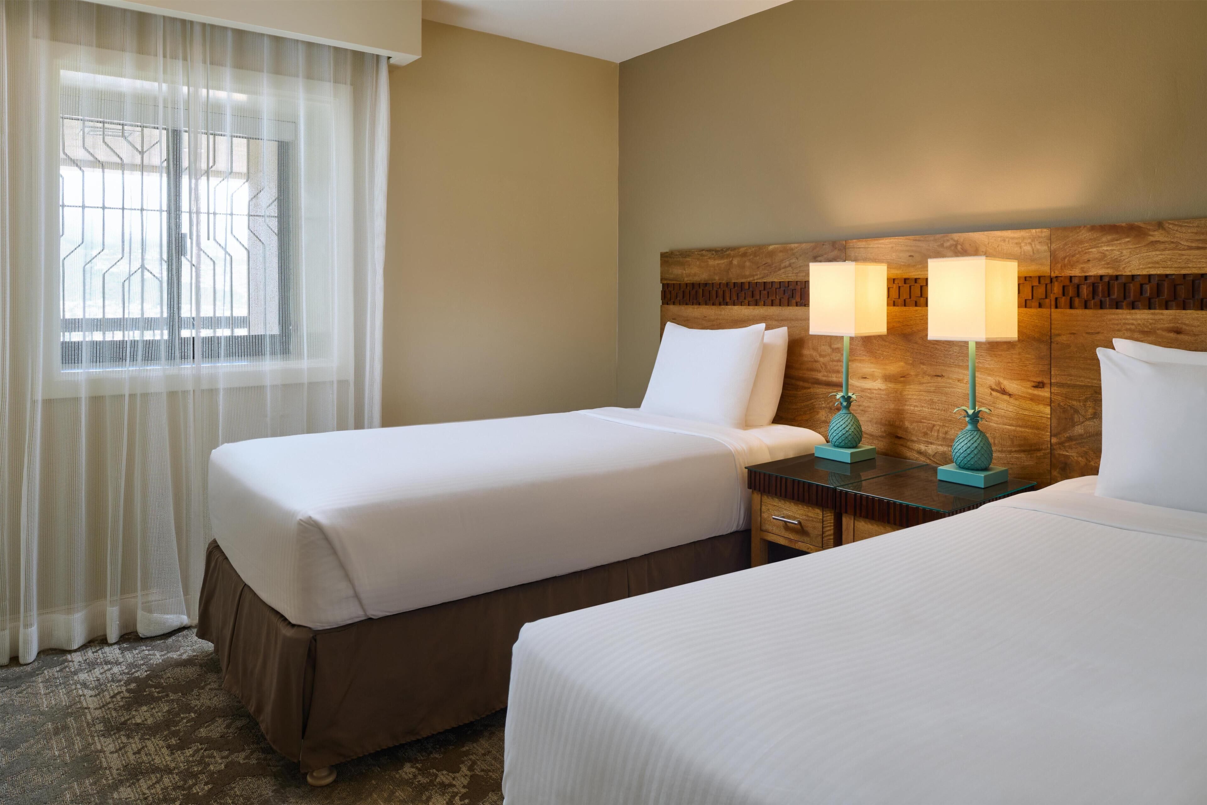 Habitación de hotel con dos camas, cabecera de madera, mesita de noche con lámparas y ventana con cortinas translúcidas.