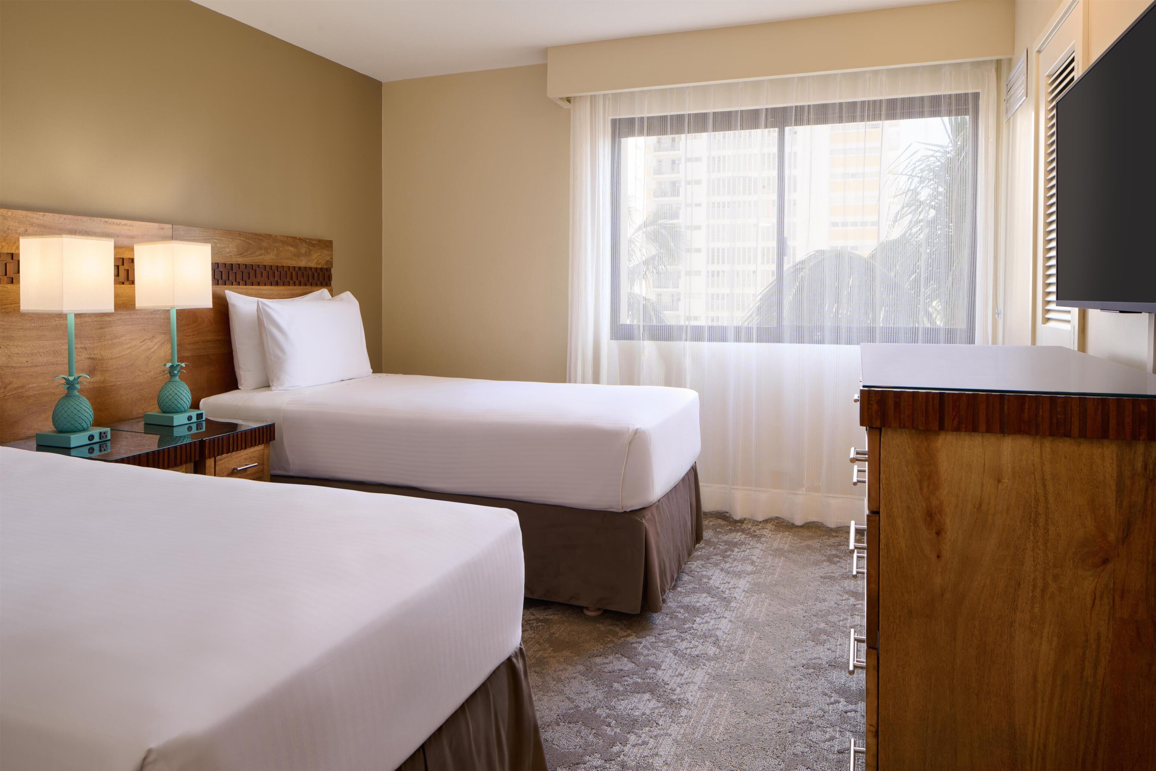 Habitación de hotel con dos camas, un escritorio con lámpara, televisión y ventana con cortinas traslúcidas.