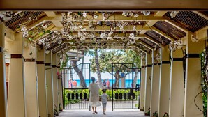 A covered walkway with a tiled floor, flanked by columns and plants, leading to a gated area with a view of palm trees and the ocean. - Aston Waikiki Beach Tower (Honolulu)