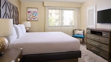 Quarto de hotel com uma cama grande, televisão, penteadeira, cadeira e janela com cortinas.