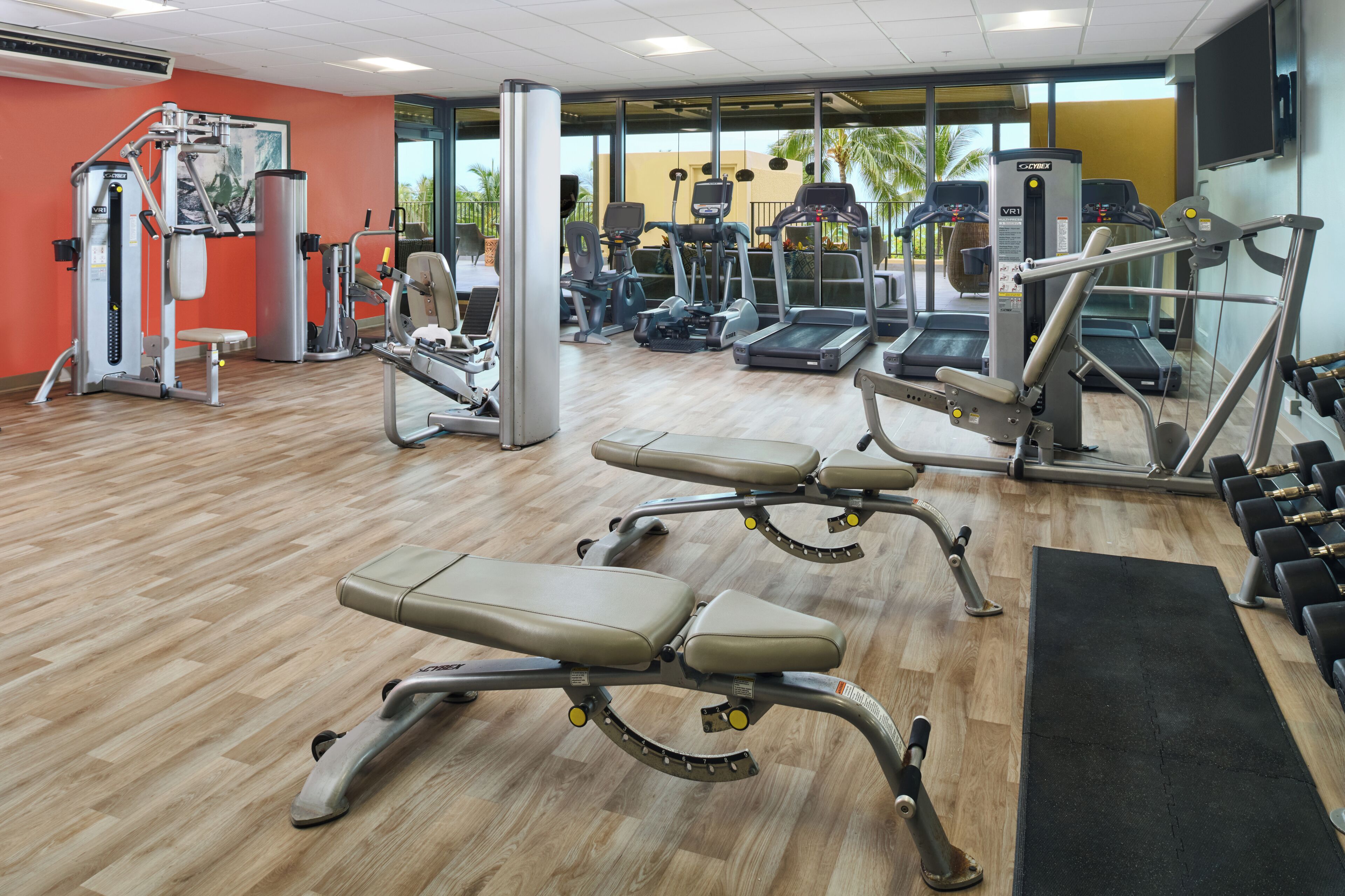 Un gimnasio con equipo de ejercicio, piso de madera y un ventanal con vistas a palmeras.
