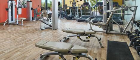 Un gimnasio con equipo de ejercicio, piso de madera y un ventanal con vistas a palmeras.