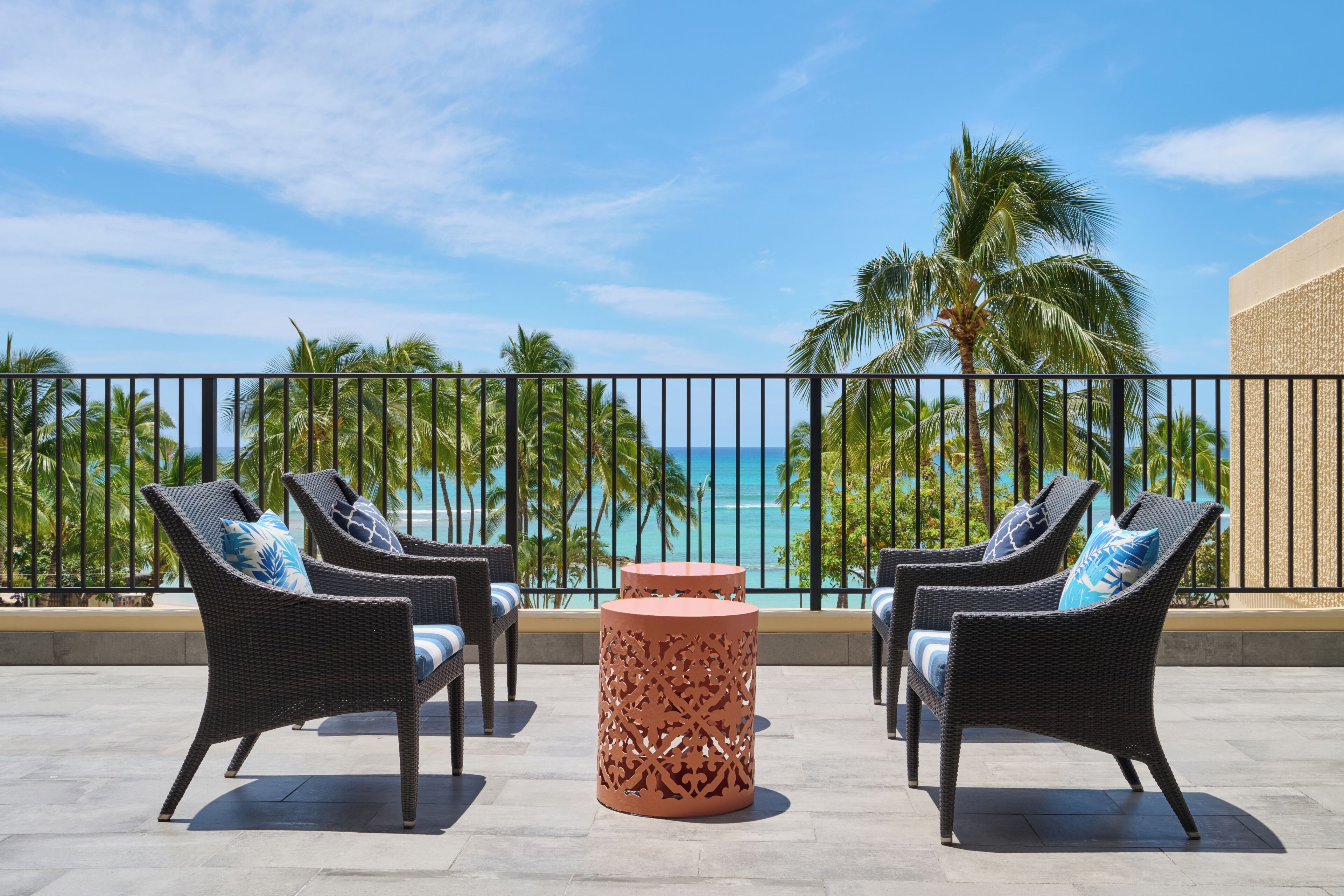 A balcony with wicker furniture and a decorative table, overlooking a beach with palm trees and a clear sky.