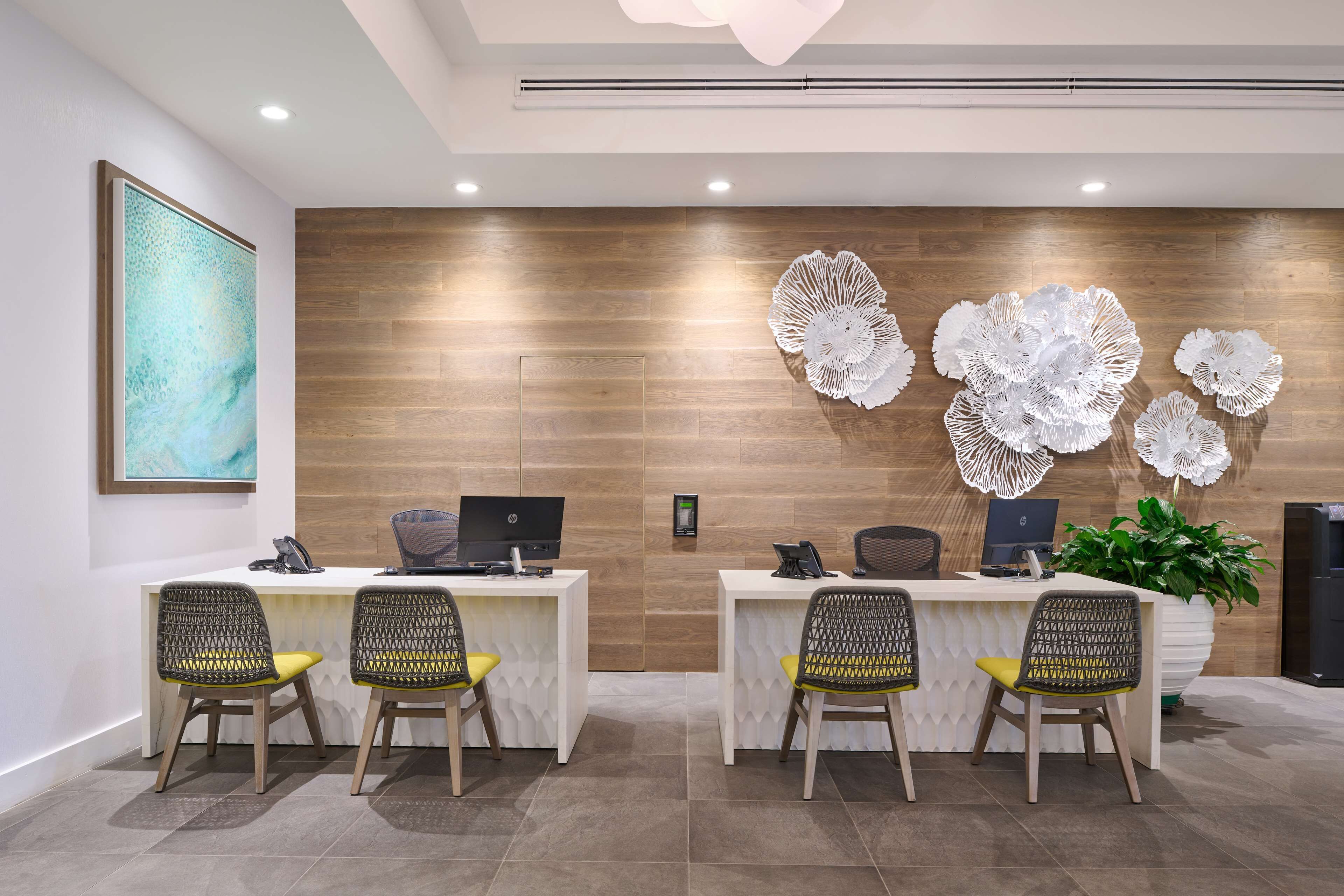 A reception area with two desks, each with a computer and a phone. There are wicker chairs with yellow cushions. A large abstract painting is on the wall, and a potted plant is on the right.