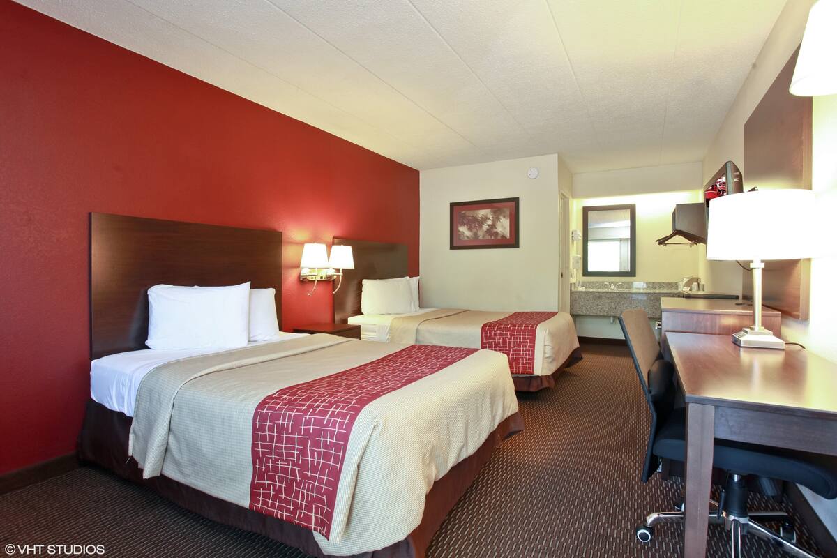 A hotel room with two beds, a desk, a chair, a mirror, and a bathroom visible in the background.