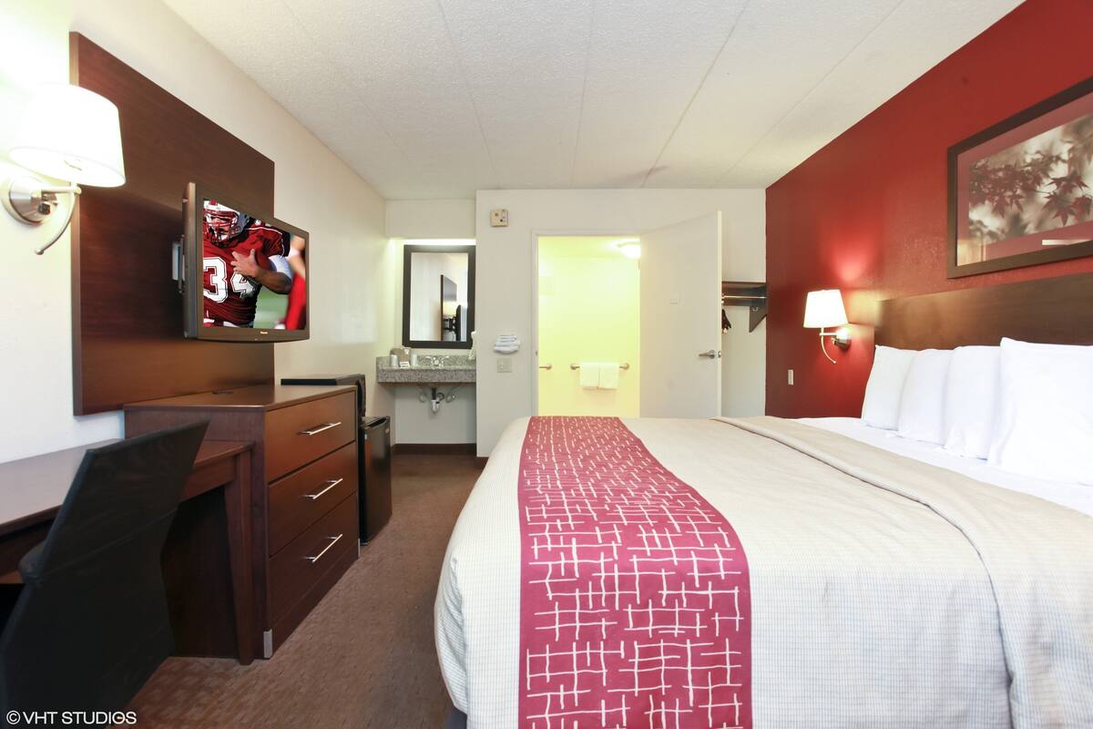 A hotel room with a bed, a desk, a TV, a mirror, and a bathroom visible through an open door.