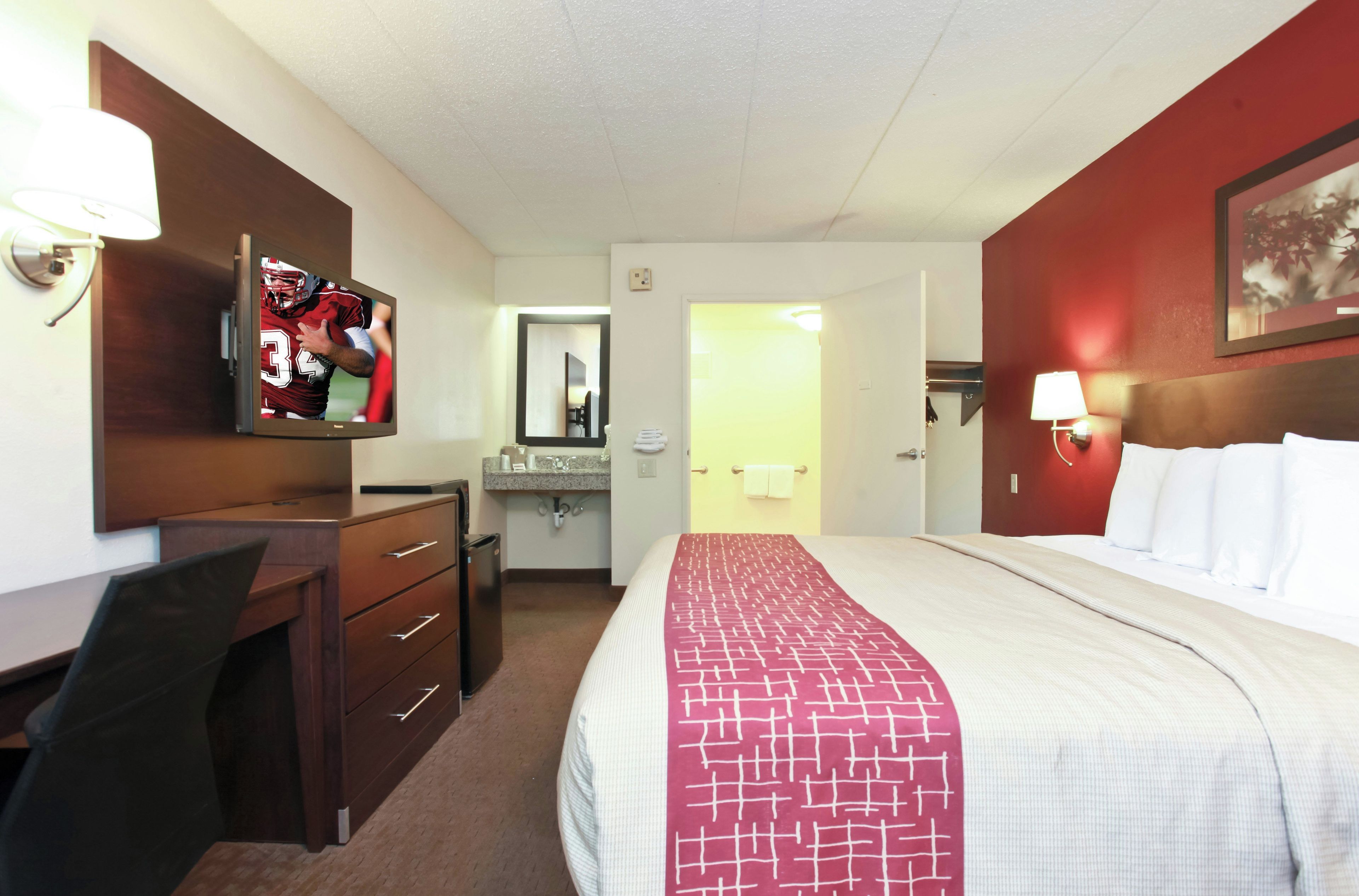 Una habitación de hotel con cama, escritorio, televisor, espejo y baño.