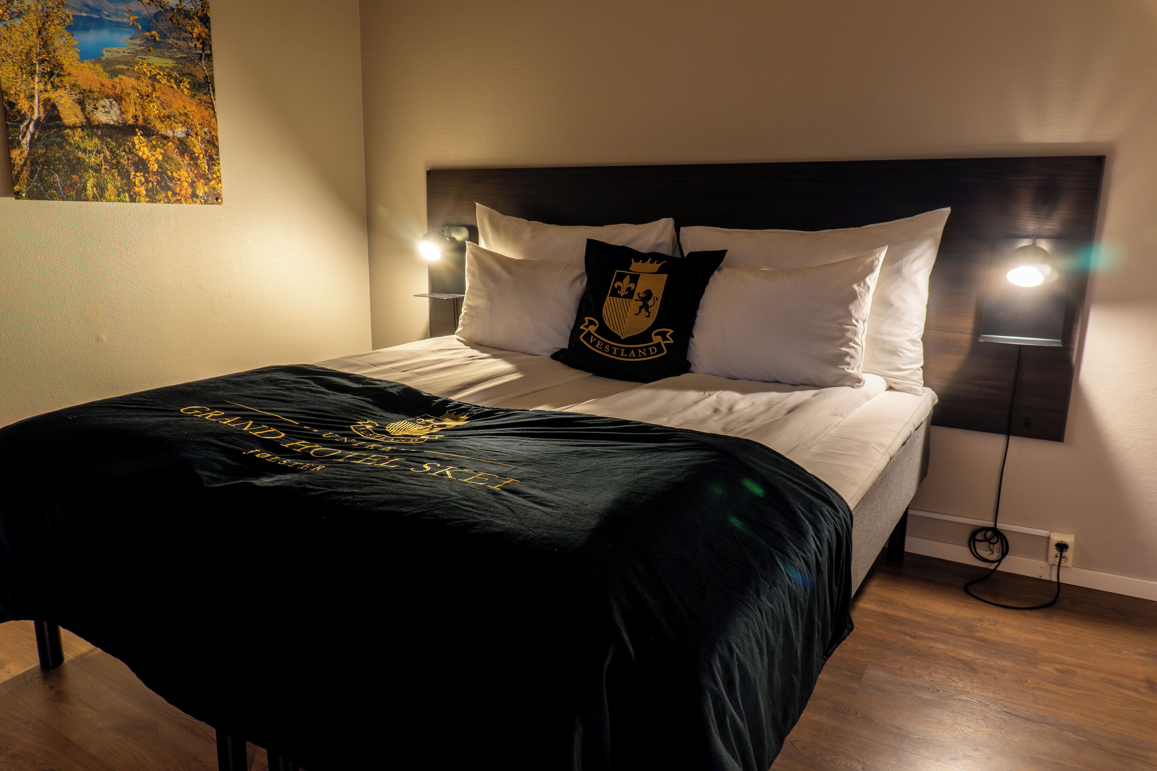 A neatly made bed with a black bedspread and a pillow with a crest. There are two bedside lamps and a painting on the wall.