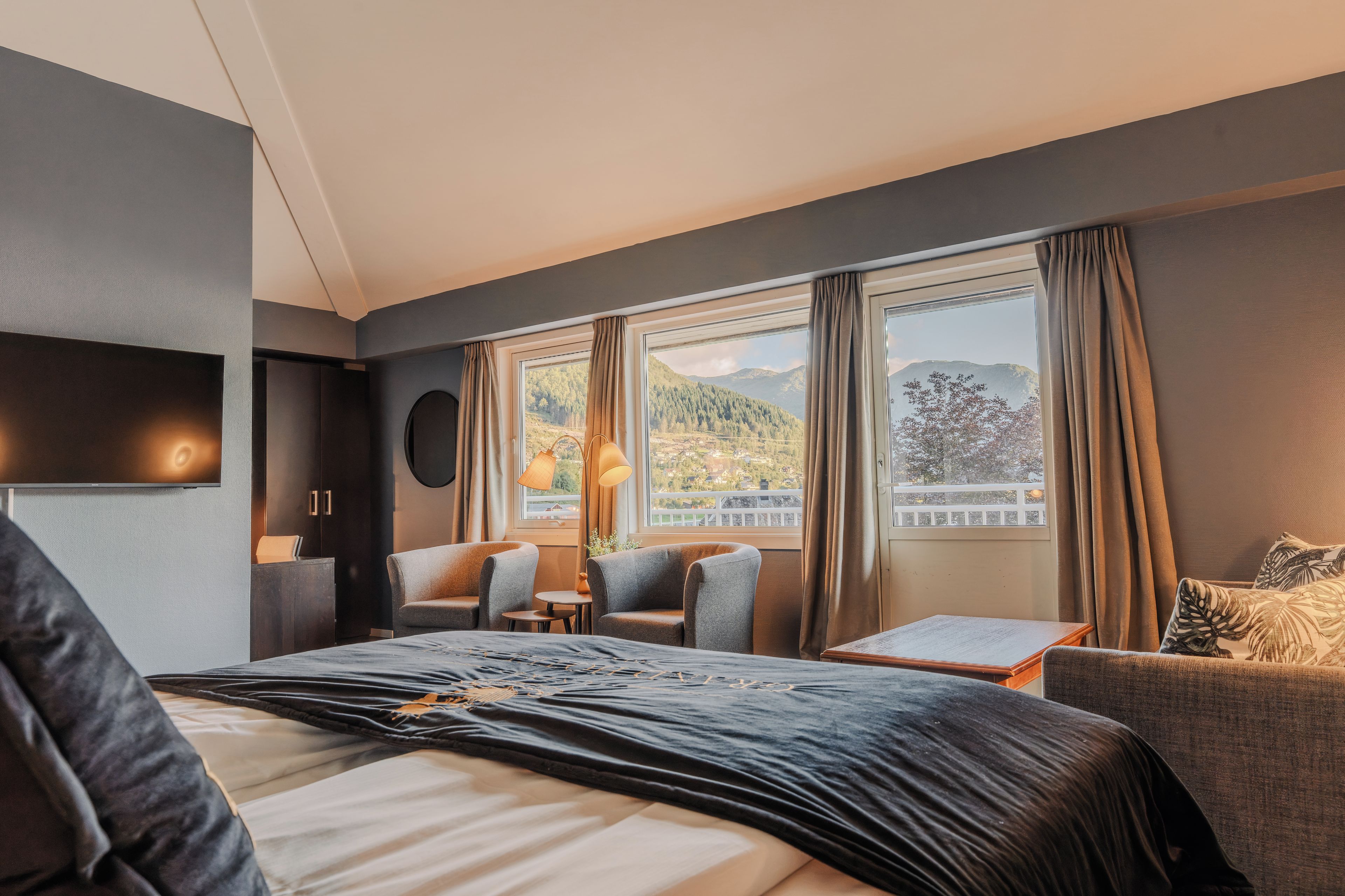 Un cuarto de hotel moderno con una cama grande, dos sillones, una mesita y vistas a un paisaje montañoso a través de las ventanas.