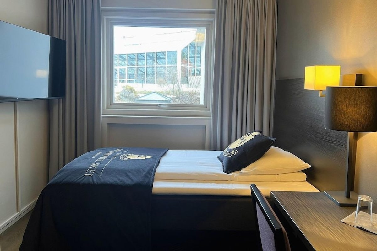 Una habitación de hotel con cama, televisión, ventana con cortinas y mesita de noche con lámpara.