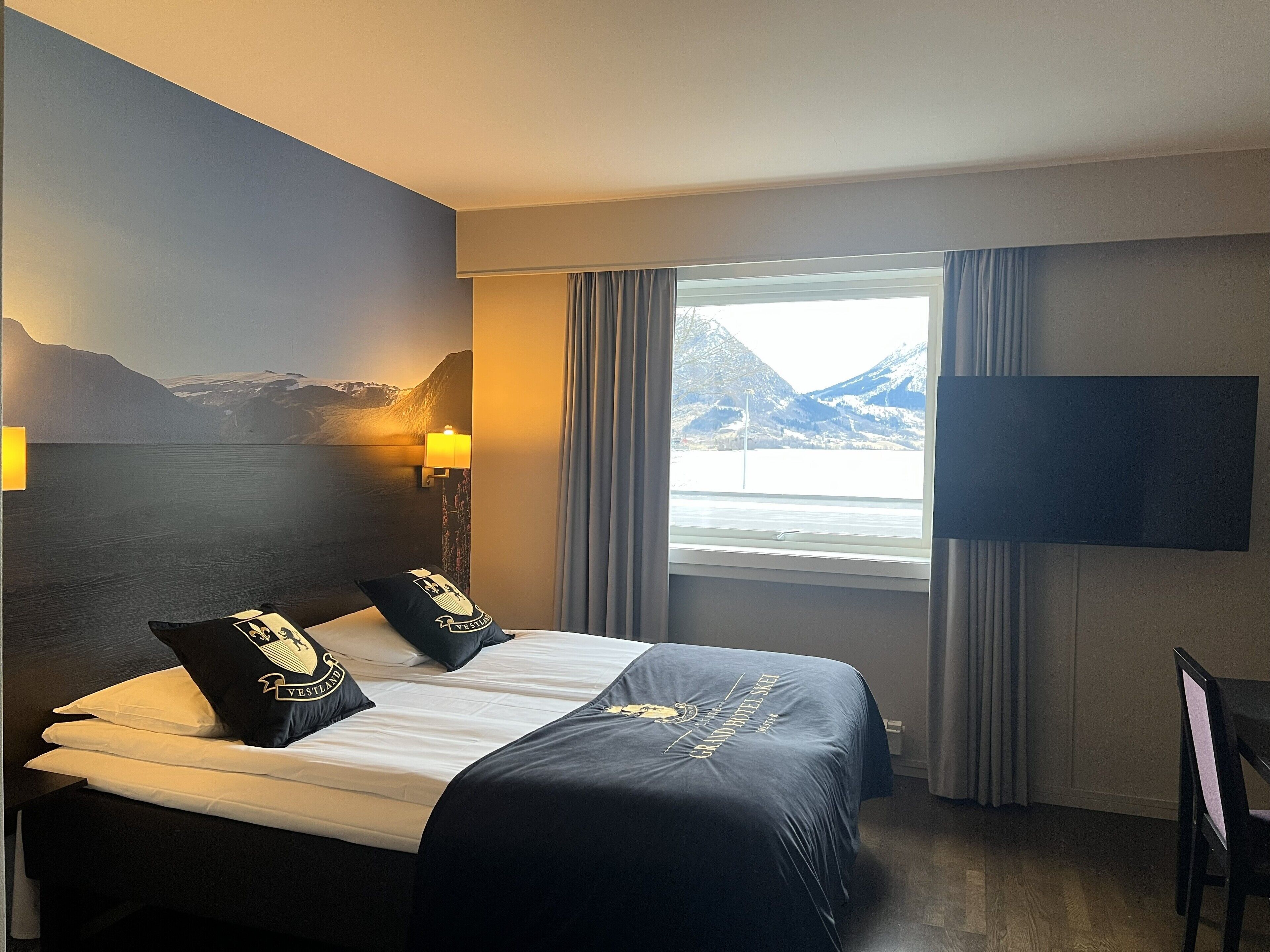 A hotel room with a bed, a TV, a window with curtains, and a wall with a mountain painting.