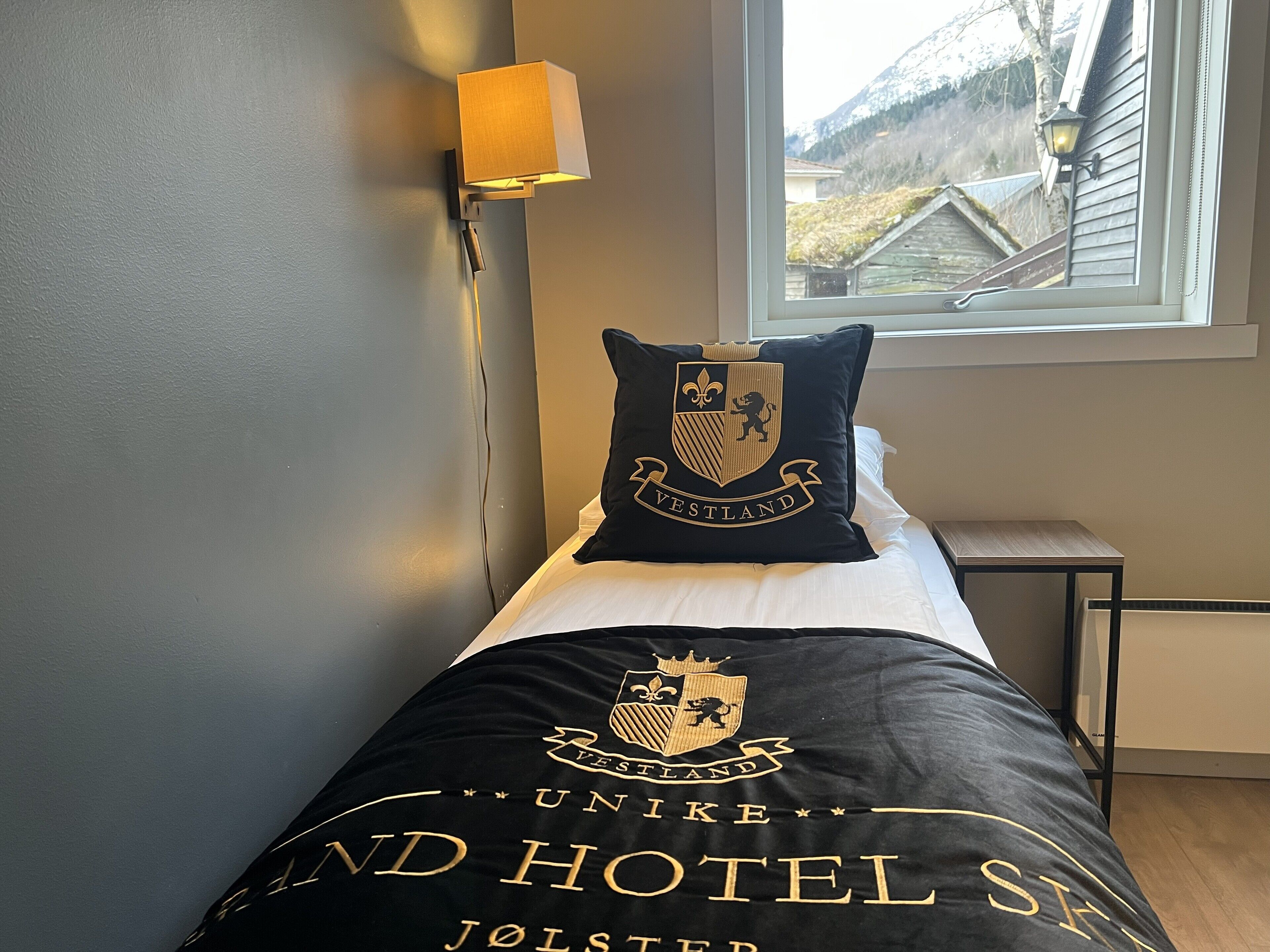 Een bed met een zwart sprei waarop het logo en de naam van Hotel Unike staan, een kussen met een familiewapen, en een raam met uitzicht op een huis en bergen.