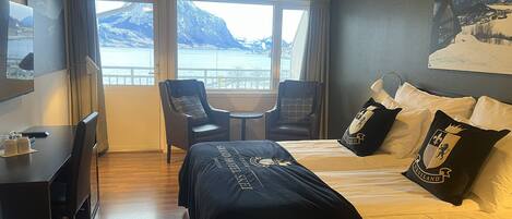 A hotel room with a bed, two chairs, a small table, a desk, and a large window offering a view of mountains and a body of water.