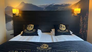 Habitación de hotel con cama, dos almohadas con escudo, un mural en la pared y dos lámparas fijadas a la pared.
