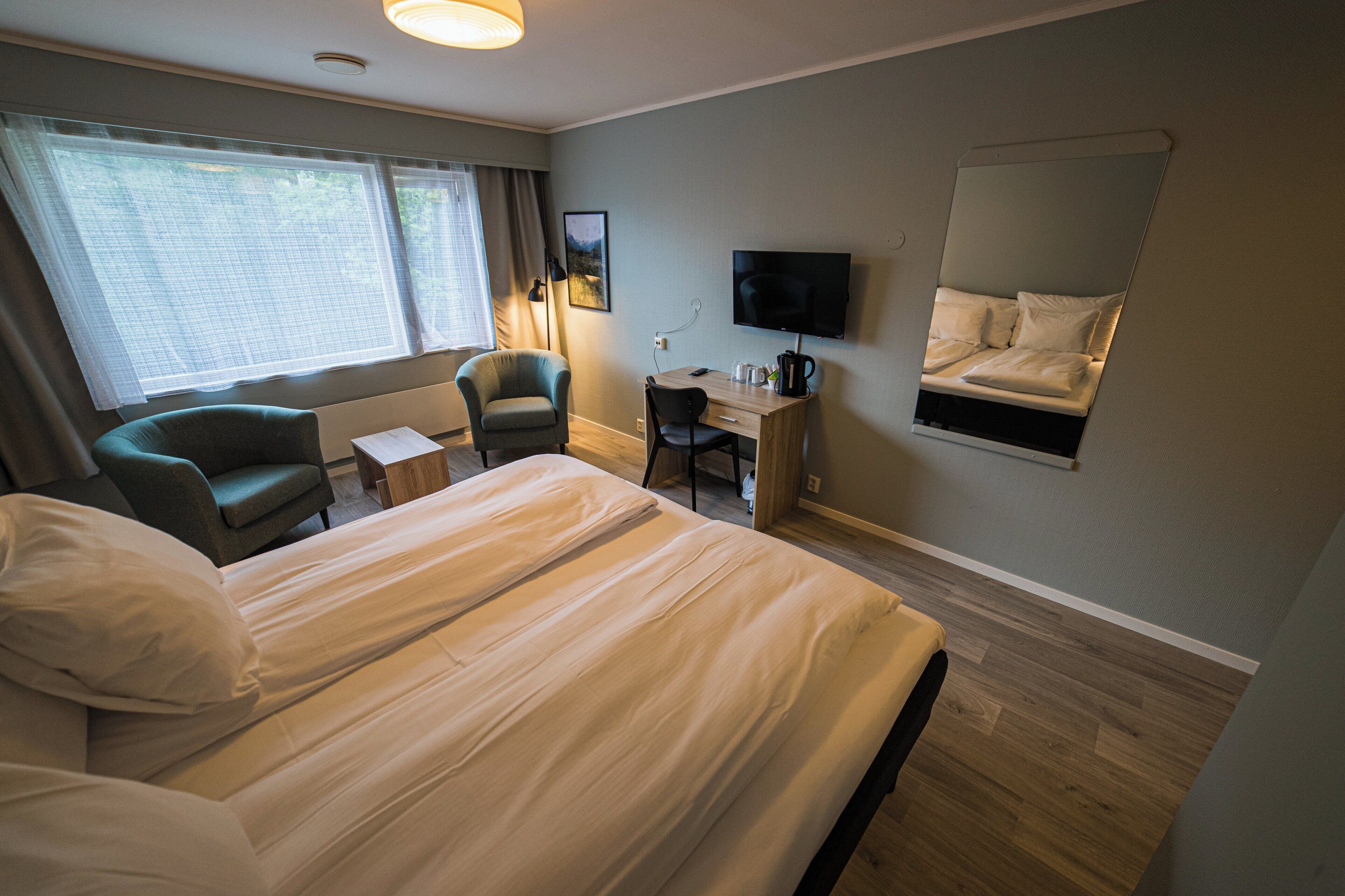 A hotel room with a bed, two chairs, a desk, a television, and a large window.