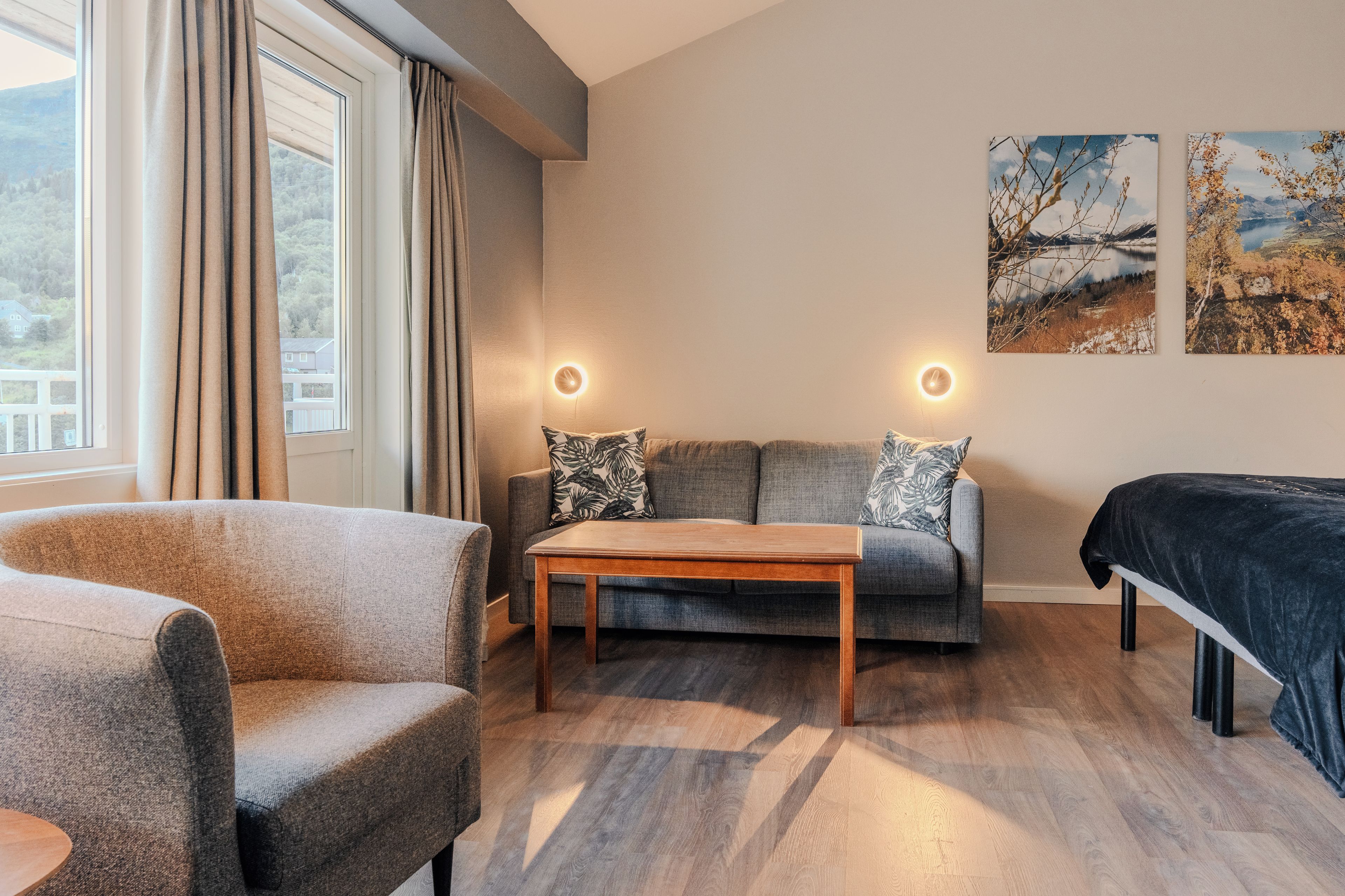 A modern living room with a sofa, armchair, and a wooden coffee table. There are two wall-mounted lights and two paintings on the wall.