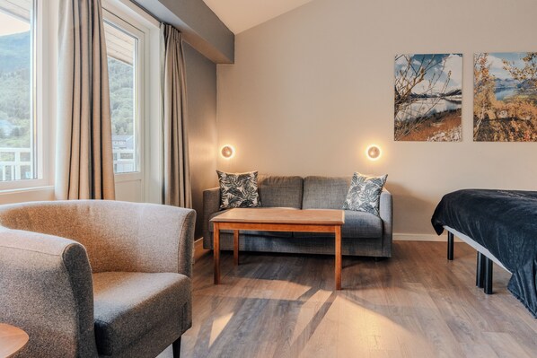 A modern living room with a sofa, armchair, and a wooden coffee table. There are two wall-mounted lights and two paintings on the wall.