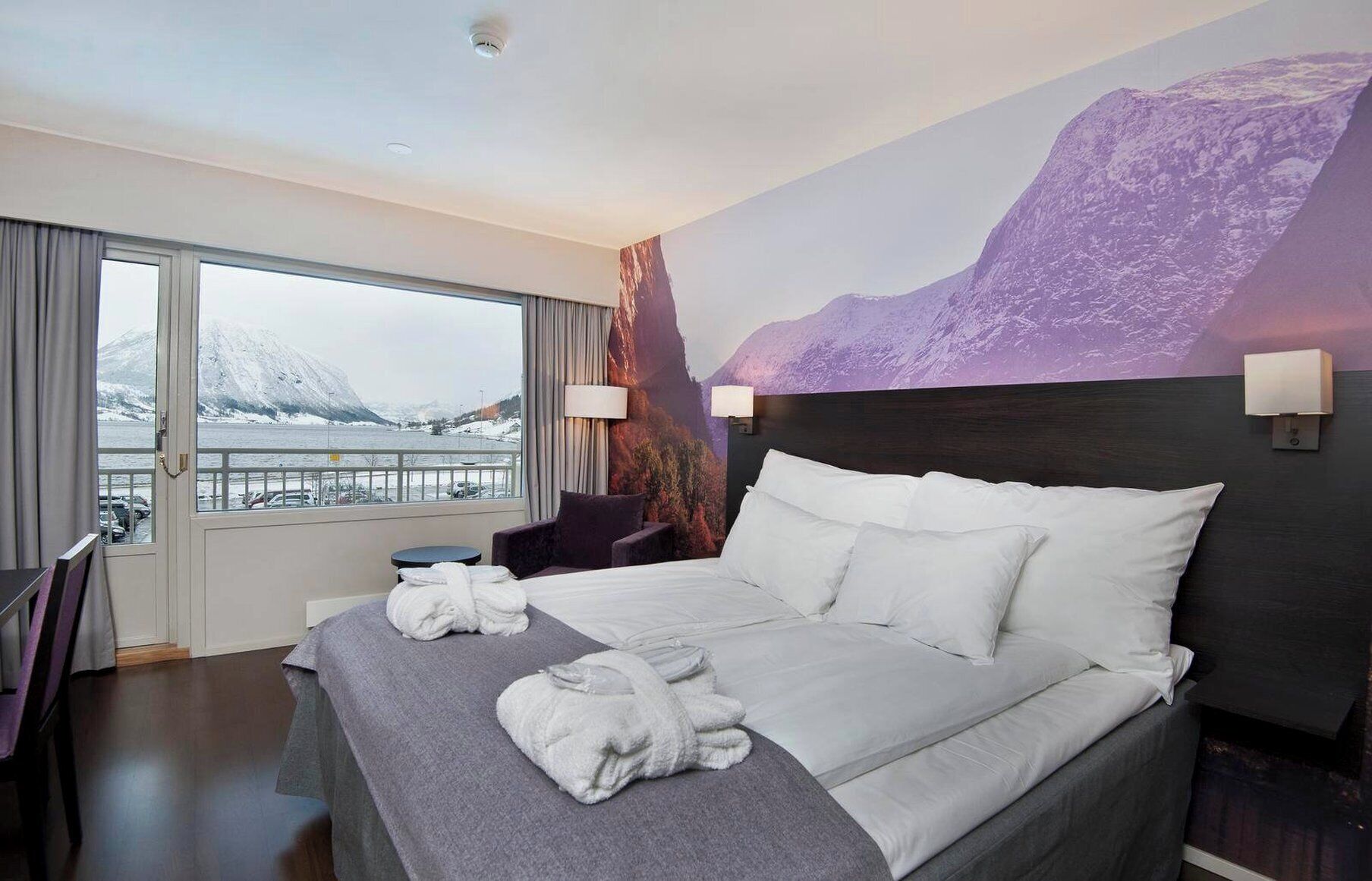 A hotel room with a bed, two pillows, a window with a view of mountains, a chair, and a table.