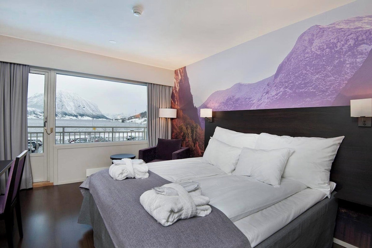 Habitación de hotel con una cama, dos almohadas, una ventana con vistas a las montañas, una silla y una mesa.