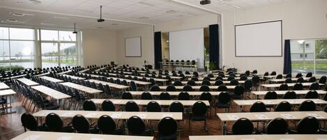 A large conference room with rows of tables and chairs, large windows, and a projector screen.