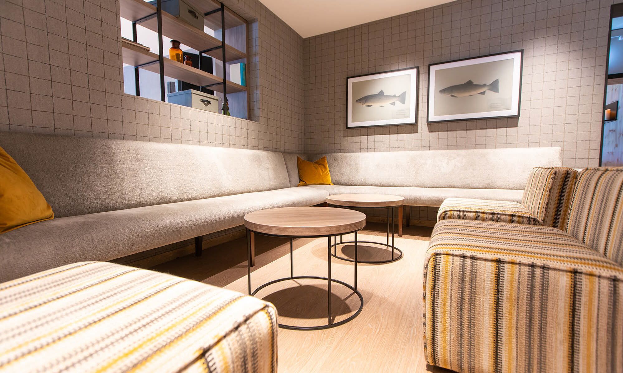 A modern living room with a sectional sofa, two round tables, and striped armchairs. There are framed fish paintings on the wall and a bookshelf with various items.