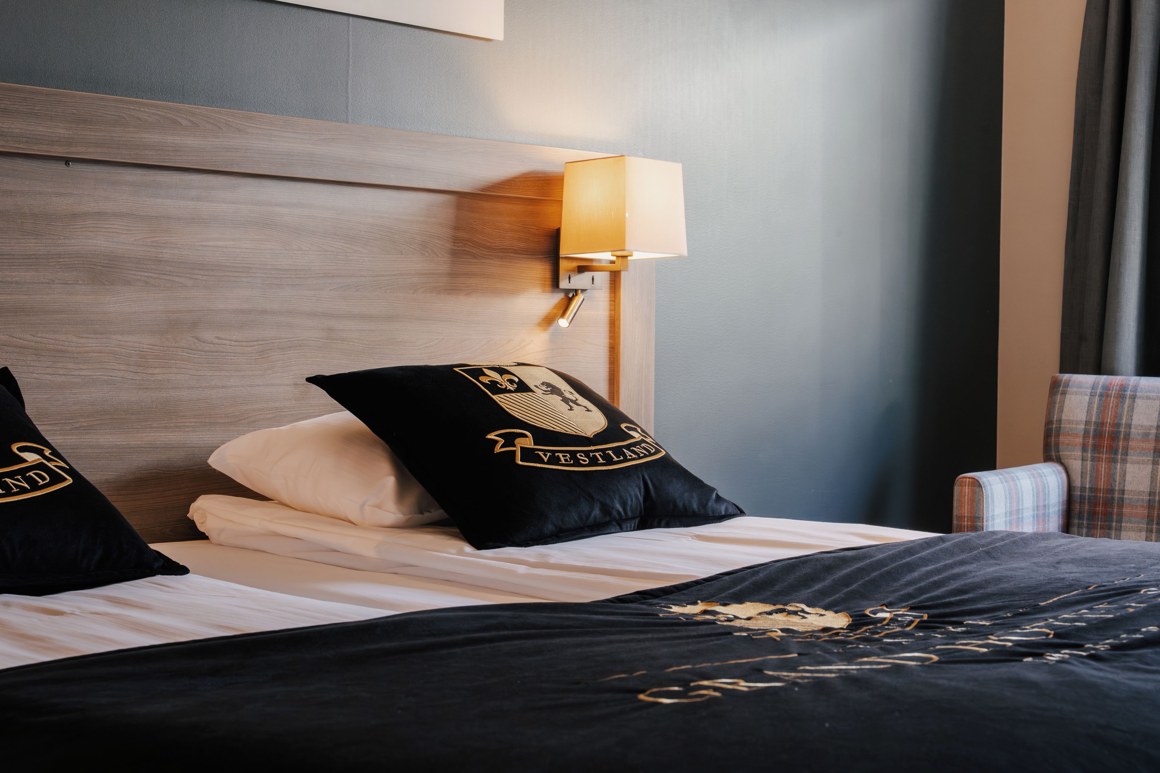 Une chambre d’hôtel avec une tête de lit en bois, une lampe de chevet et deux coussins noirs ornés de broderies dorées.