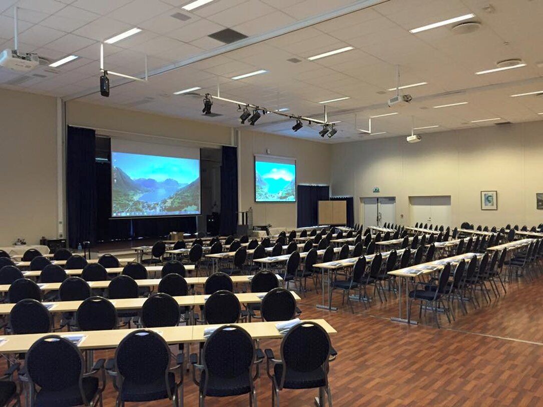 Un salón de conferencias con filas de mesas y sillas, una pantalla grande y un proyector.