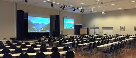 Un salón de conferencias con filas de mesas y sillas, una pantalla grande y un proyector.