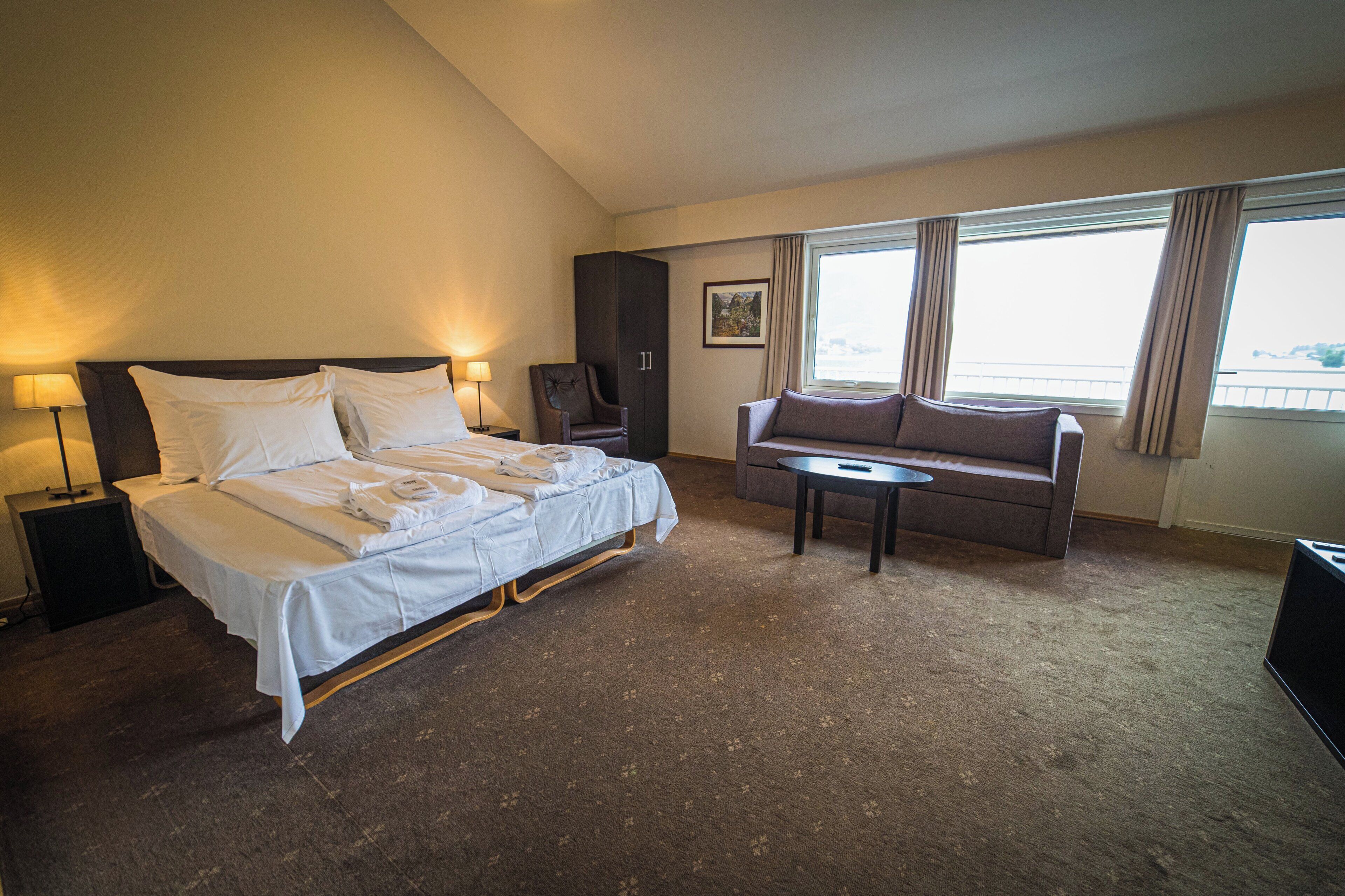 Habitación de hotel con cama, sofá, mesita, silla, mesita de noche y ventana con cortinas.