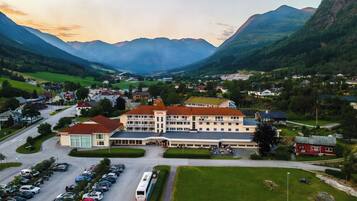 Un hotel con un edificio grande, estacionamiento y montañas al fondo.