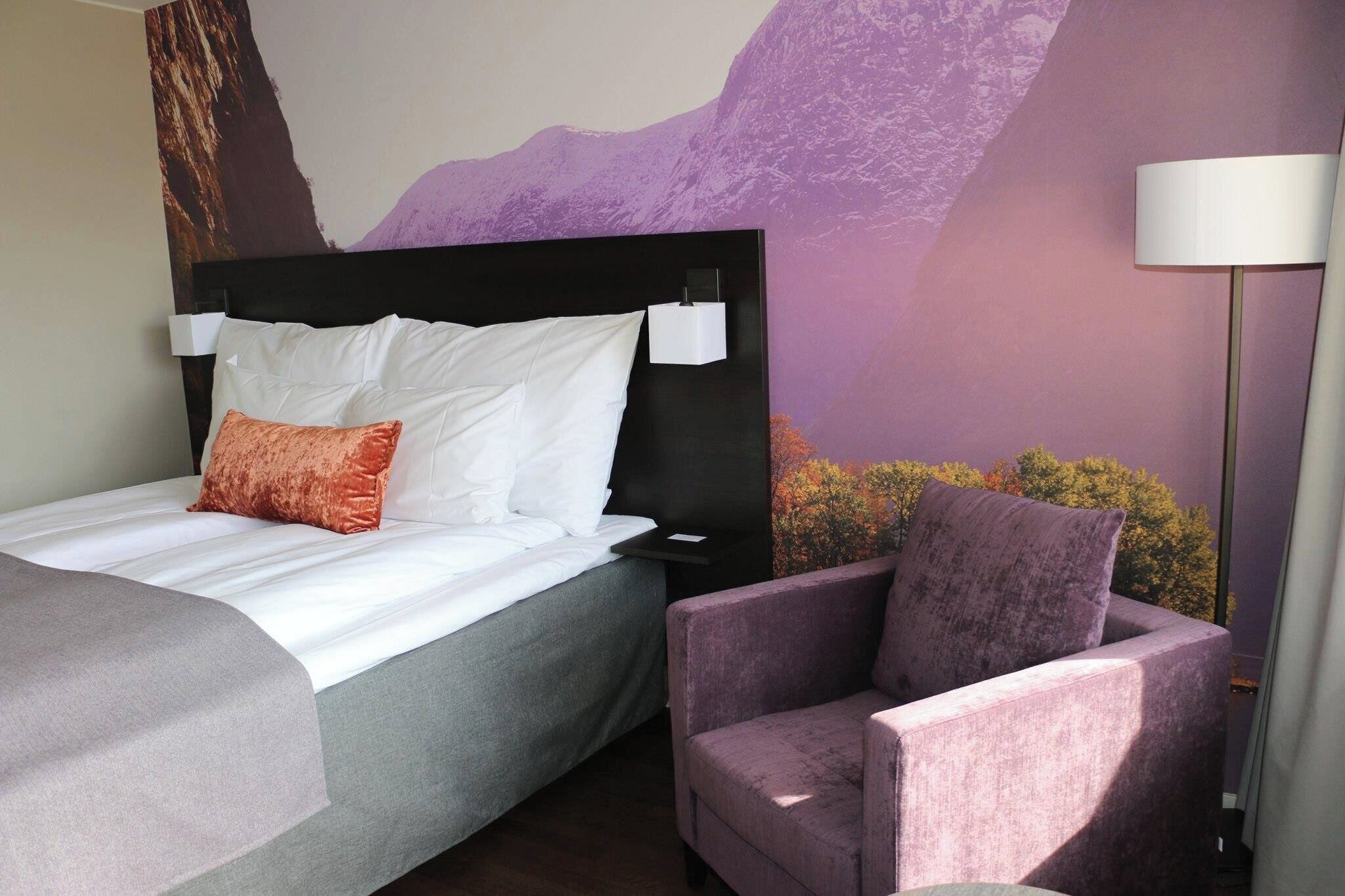 Quarto de hotel com cama, uma cadeira, mesa de cabeceira e um mural na parede.
