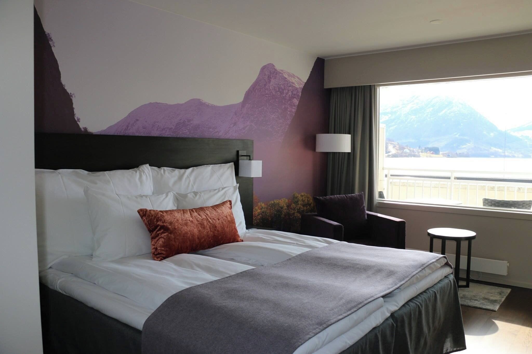 Habitación de hotel con cama, una silla, una mesita, una lámpara y ventana con vista a la montaña.