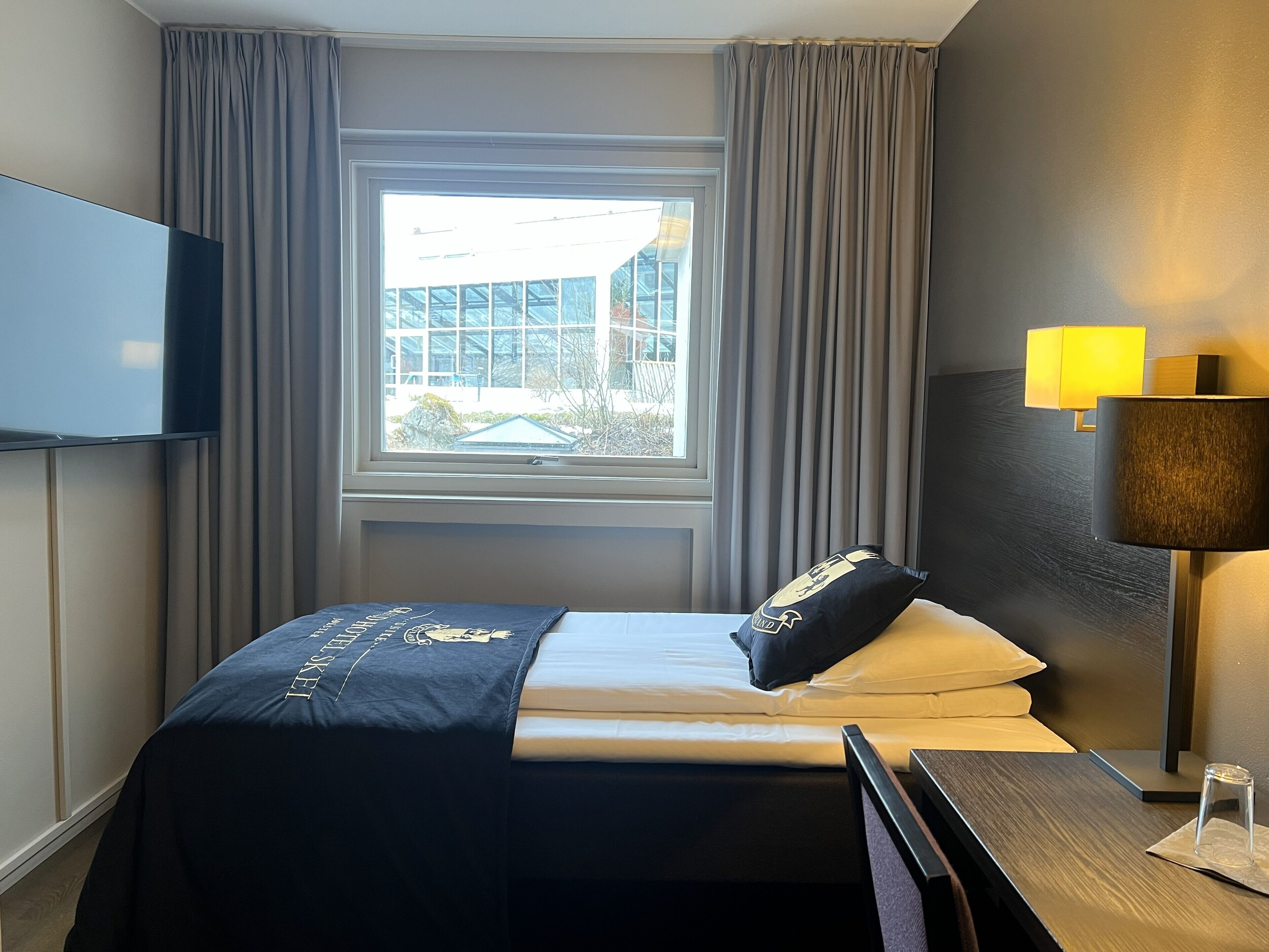 Ein Hotelzimmer mit einem Bett, einem an der Wand befestigten Fernseher, einem Fenster mit Vorhängen und einer Lampe.