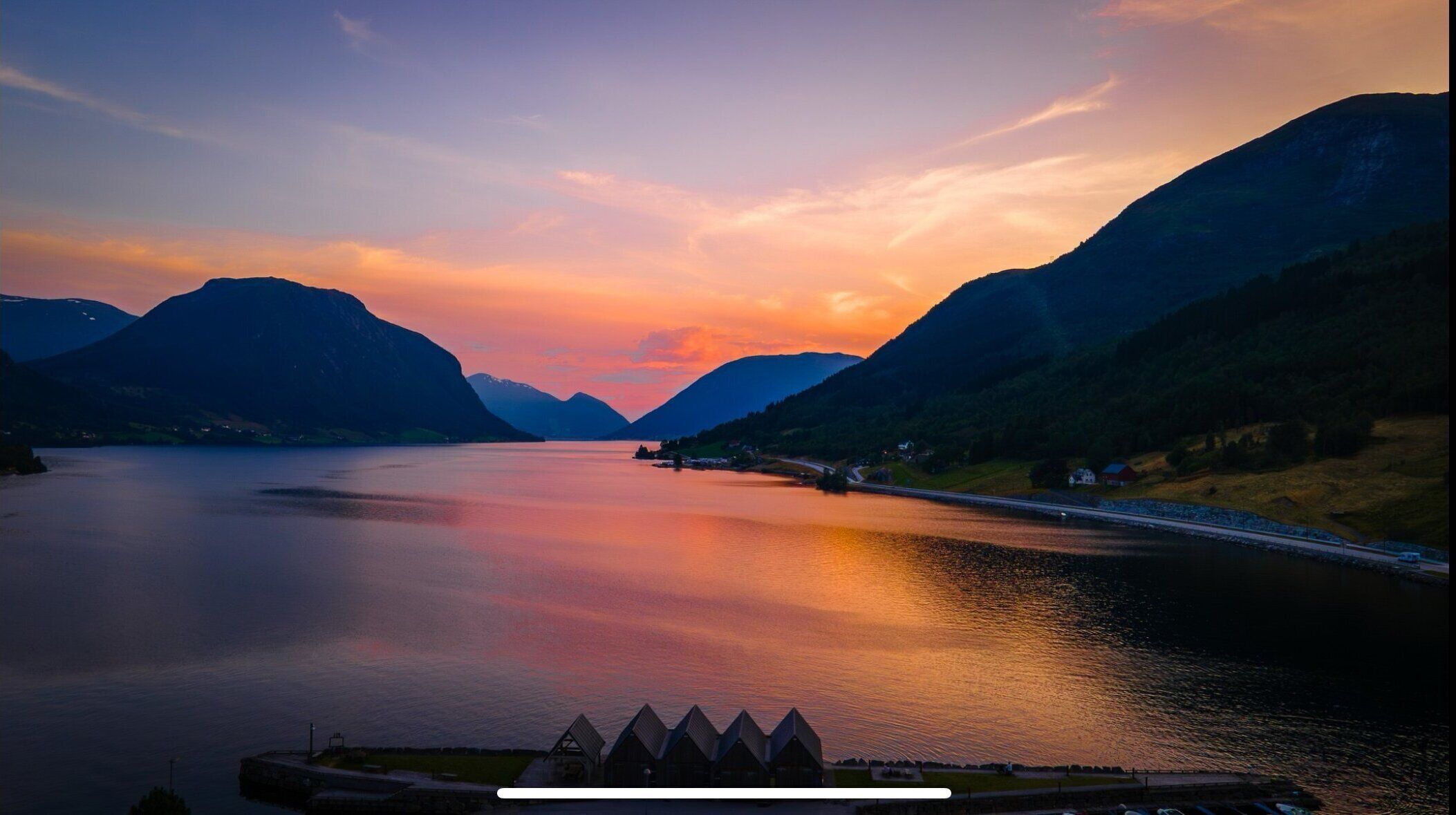 A serene lake with mountains, a road, and a row of triangular-roofed buildings.