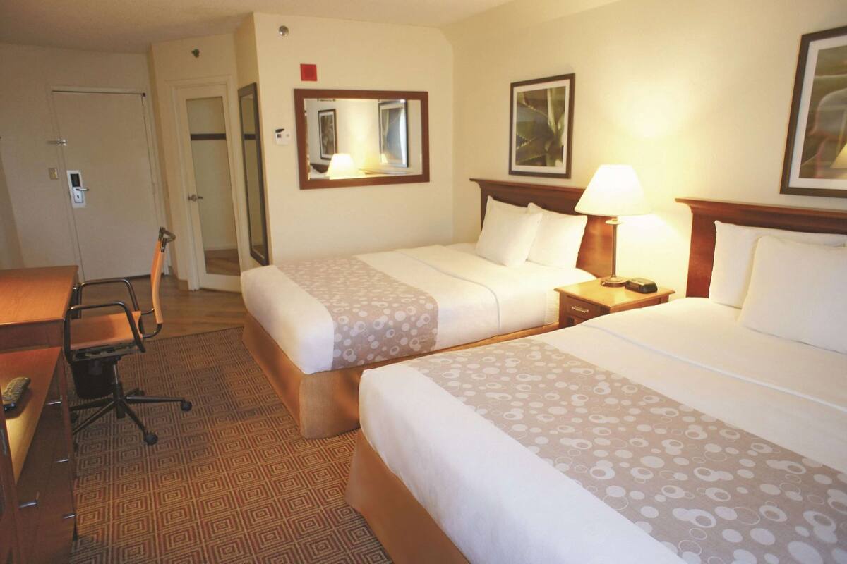 A hotel room with two beds, a desk, a chair, a lamp, a mirror, and a door.