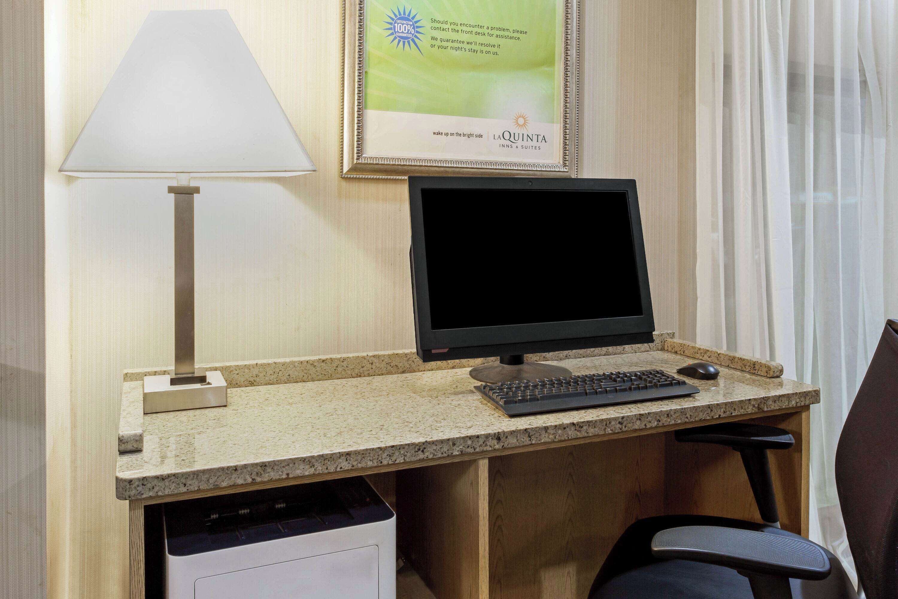 A hotel room with a desk, computer, chair, and a lamp.