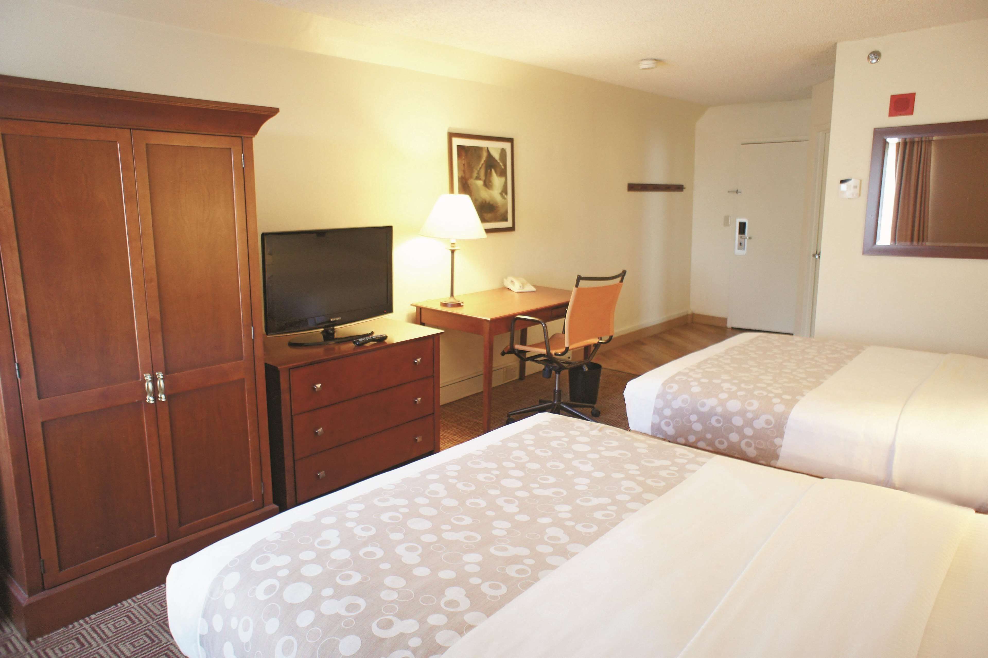A hotel room with a wooden wardrobe, a desk with a lamp, a chair, and two beds with patterned bedding.