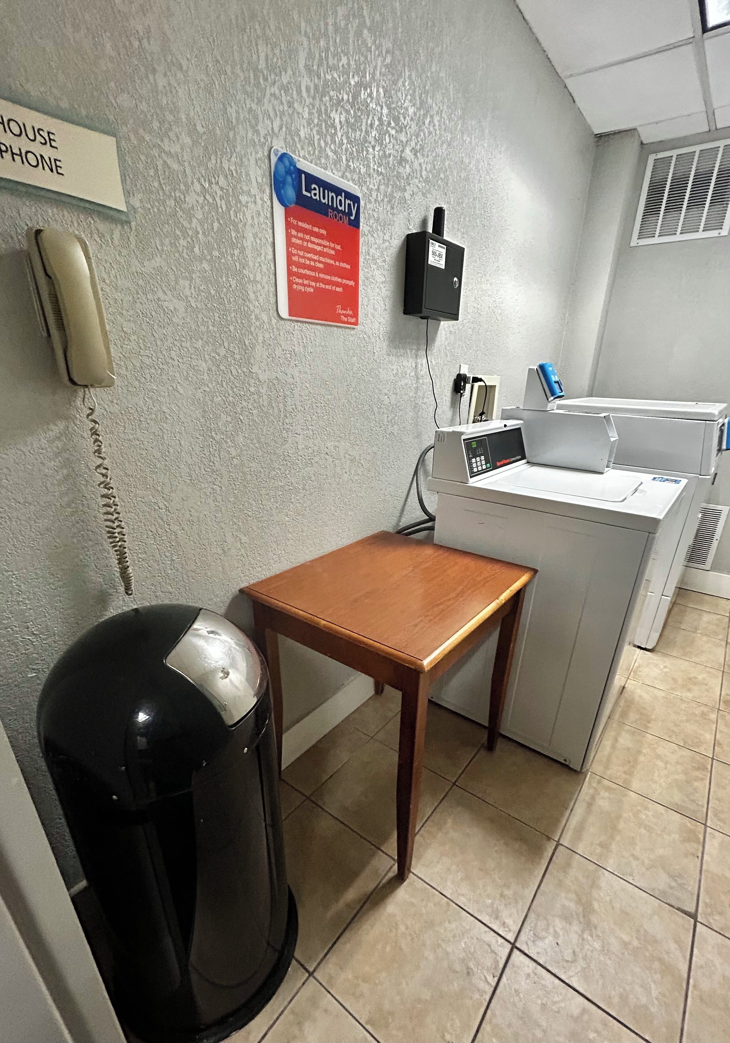 Une buanderie équipée d’un lave-linge, d’une poubelle, d’une table en bois, d’un téléphone et d’un panneau indiquant la fonction de la pièce.