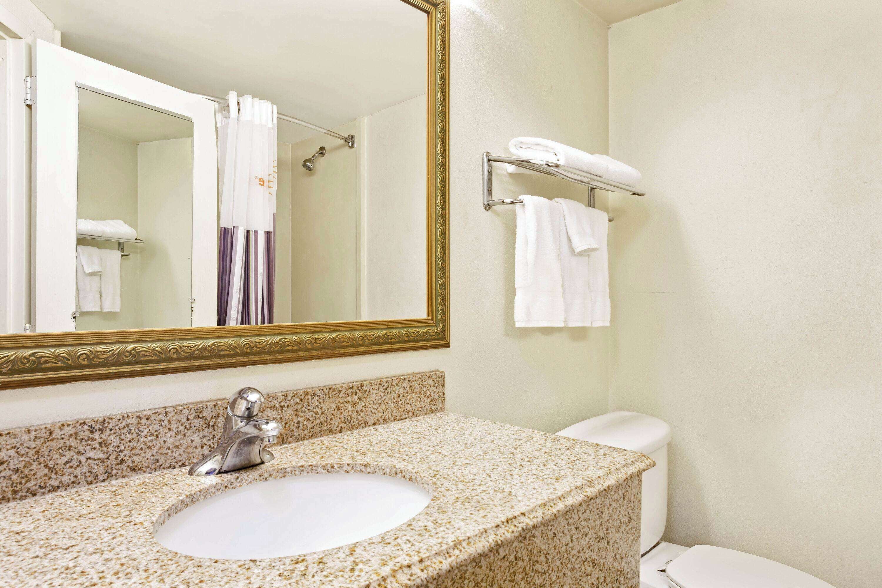 A bathroom with a granite countertop sink, a large mirror with a gold frame, and a towel rack with folded towels.