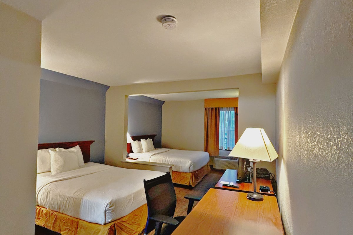 A hotel room with two beds, a desk, a chair, and a lamp.