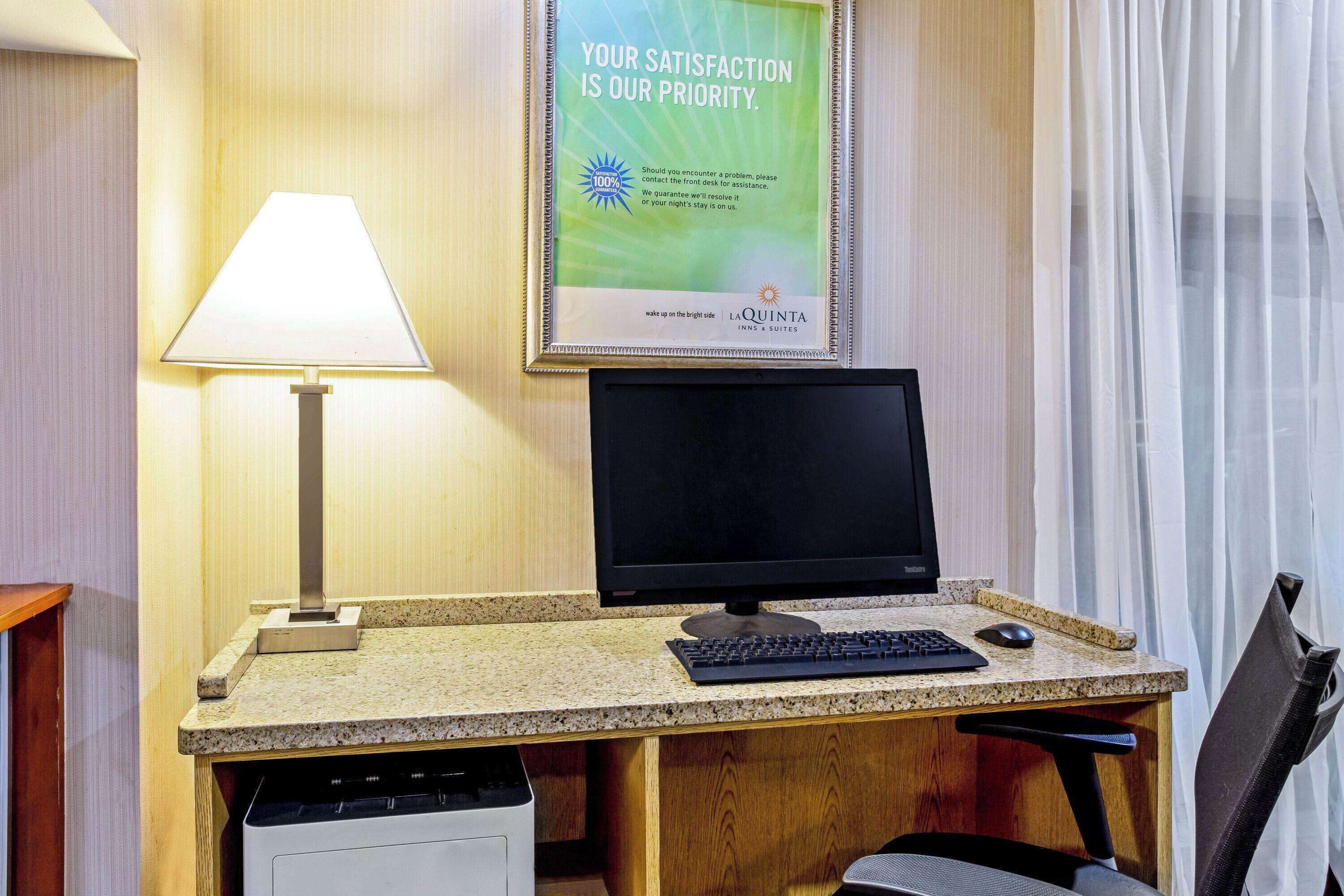 A hotel room with a desk, computer, and lamp. A poster on the wall reads "Your satisfaction is our priority."