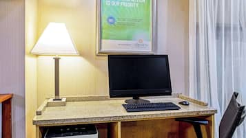 A hotel room with a desk, computer, and lamp. A poster on the wall reads "Your satisfaction is our priority."